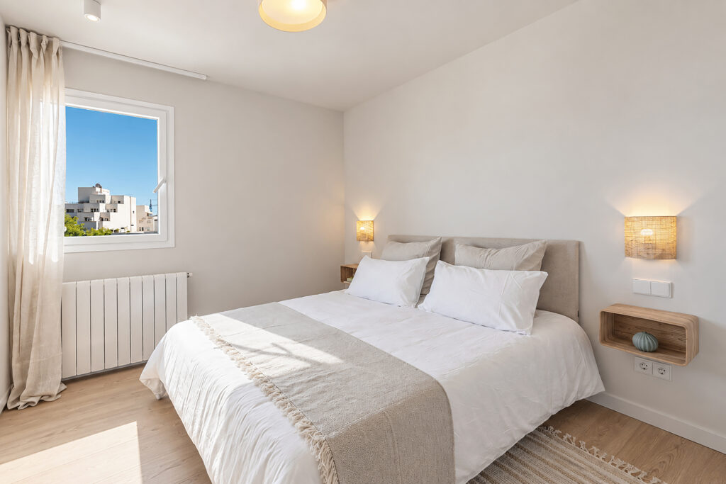 Dormitorio luminoso con pavimento de madera, ropa de cama clara y ventana exterior