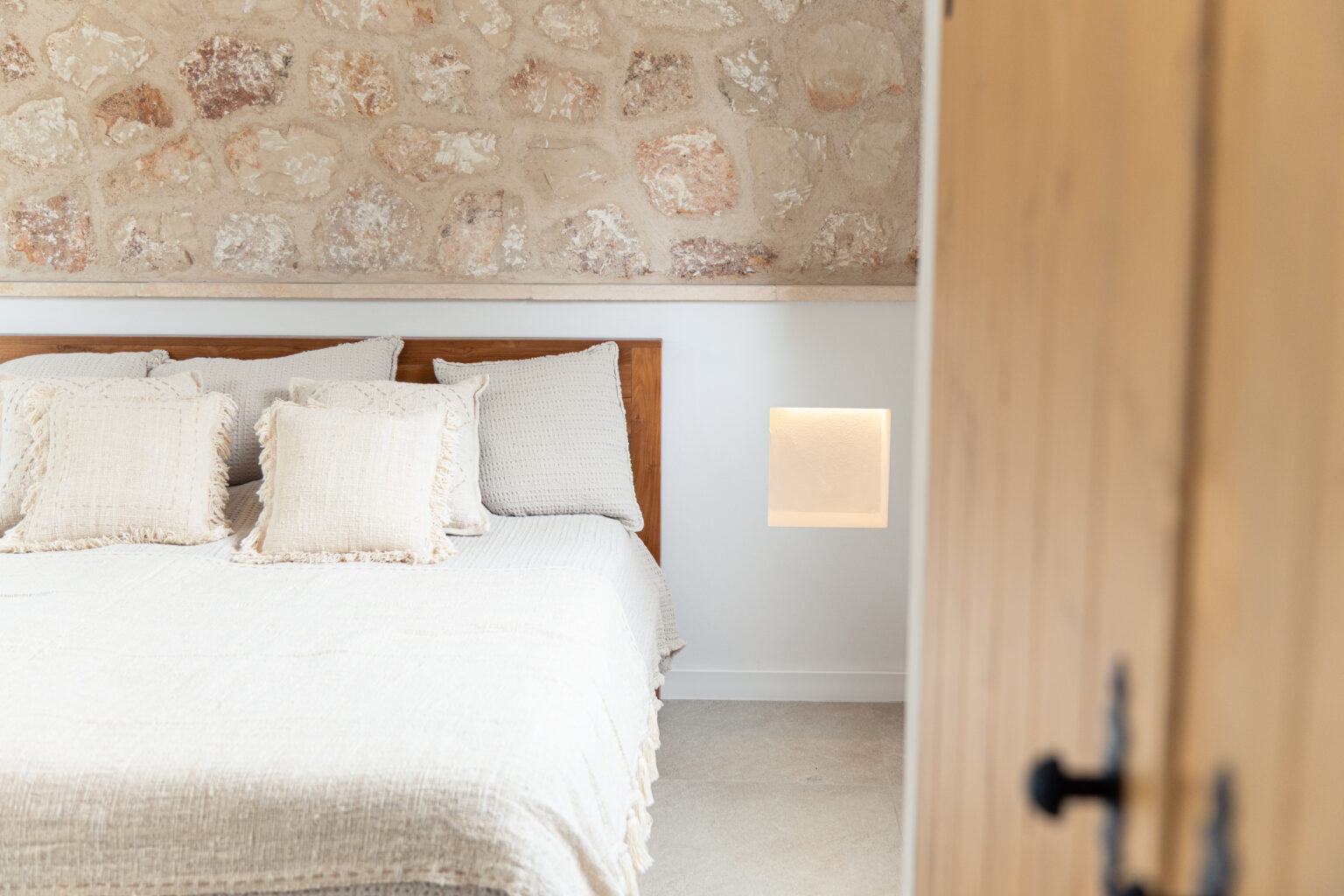 Dormitorio de S'Embat con cabecero de madera, pared de piedra y hornacinas iluminadas